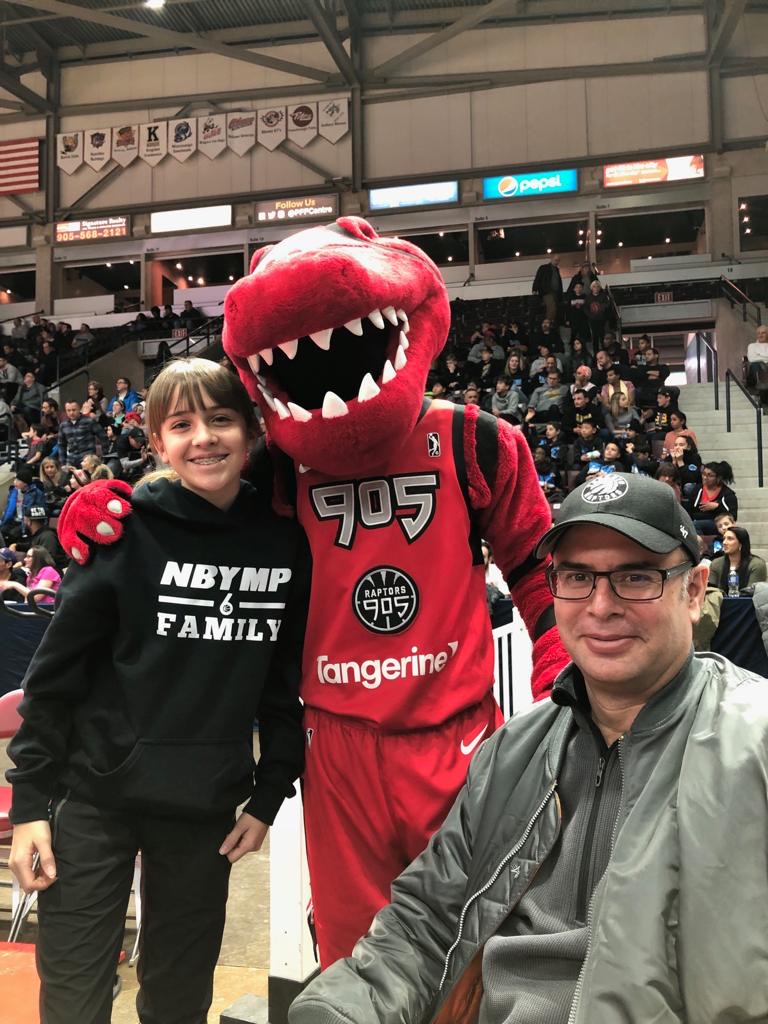 an NBYMP mentee wearing a black NBYMP hoodie stands beside the Toronto Raptor's mascot with a seated adult